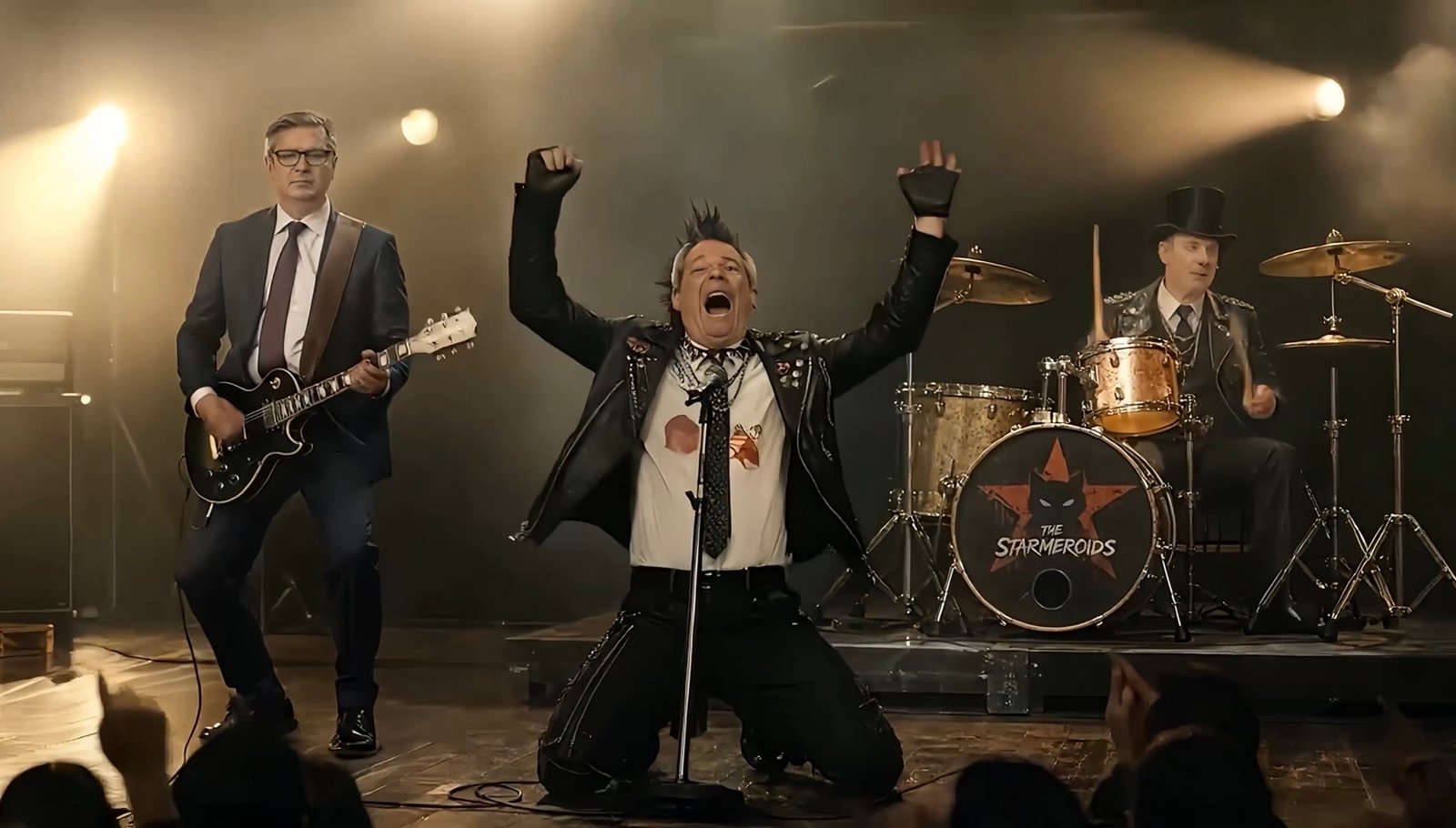 A punk singer kneels at the microphone with both arms raised while a suited guitarist performs beside him and a top-hatted drummer plays behind a Starmeroids-branded drum kit.